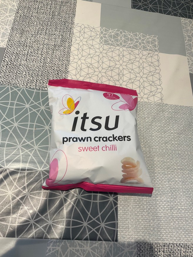 A packet of Itsu Sweet Chilli Prawn Crackers lies on a tablecloth with duck egg colour scheme patterns.