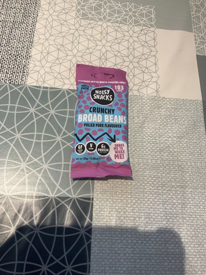 A packet of Noisy Snacks Pulled Pork Broad Beans lies on a tablecloth with various duck egg colour scheme patterns.
