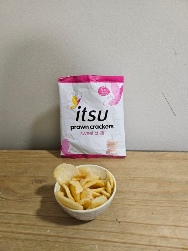 The empty packet for the Itsu Sweet Chilli Prawn Crackers sits on a light brown table and leans against a white wall. A small white bowl filled with the crackers sits in front of the packet.
