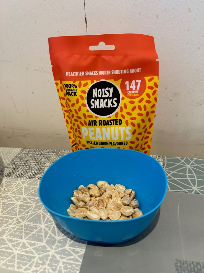 A packet of Noisy Snacks Pickled Onion Peanuts sits on a tablecloth with various duck egg colour scheme patterns and leans against a white wall. A small plastic blue bowl filled with the peanuts sits in front of the packet.