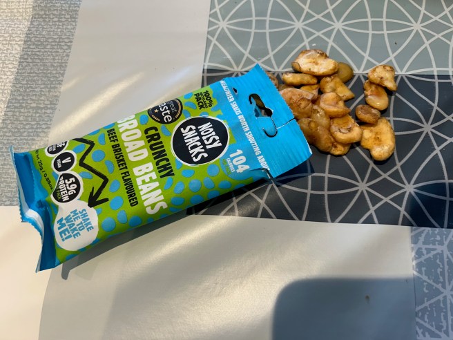 A packet of Noisy Snacks Beef Brisket Broad Beans and some of the beans themselves lie on a tablecloth with various cuck egg colour scheme patterns.