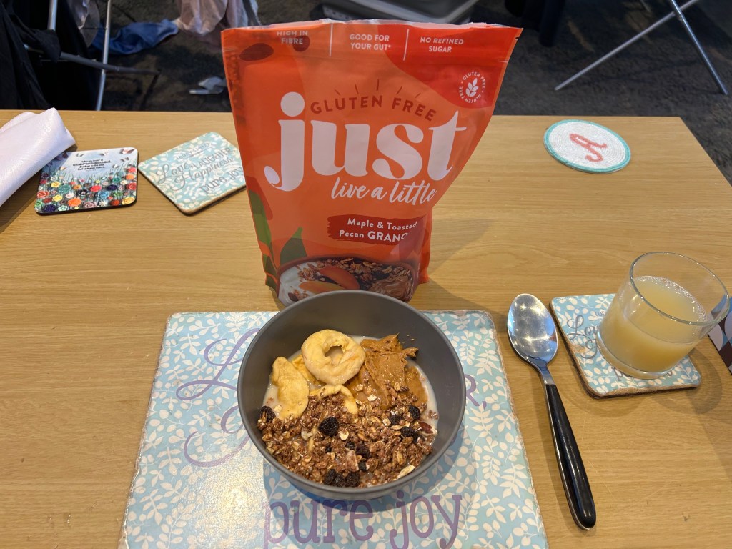 A packet of Just Live a Little granola, a grey bowl of the granola with dried fruit and peanut butter, and a glass of apple juice sit on a coffee table.