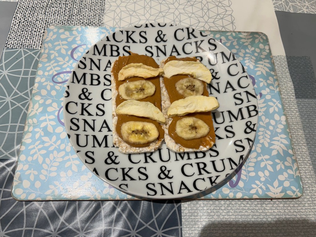 Some rectangular rice cakes with peanut butter and dried fruit on them sit on a white side plate with black text on it.