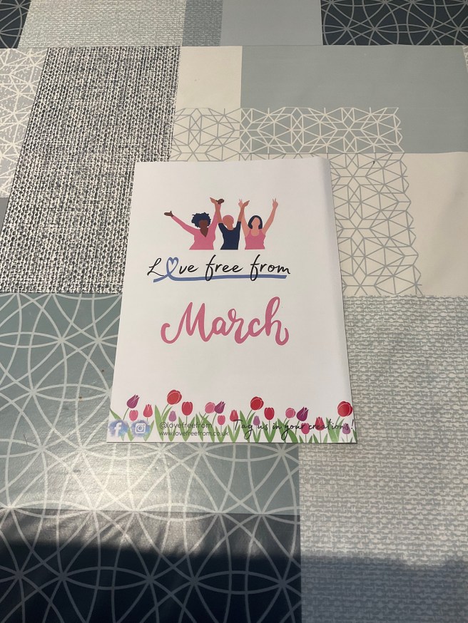 A leaflet for the March 2024 Love Free From box lies on a tablecloth with duck egg colour scheme patterns. The leaflet has a picture of 3 women near the top and a picture of some flowers at the bottom.