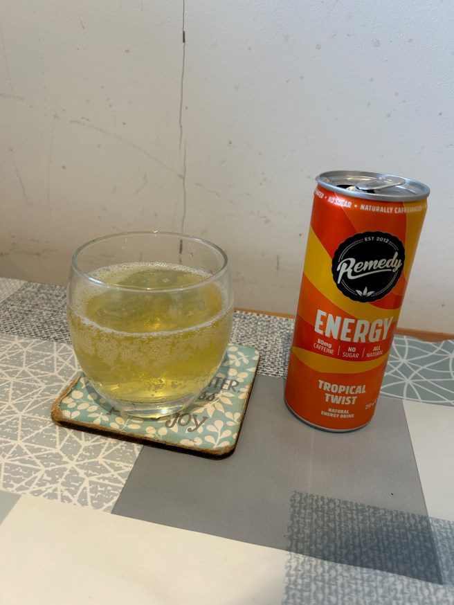 A glass of Remedy Energy drink and its can stand against a white wall and on a tablecloth with duck egg colour scheme patterns.