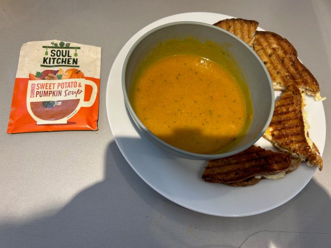 An empty packet of Soul Kitchen instant soup lies beside a plate with a bowl of the soup and a toasted cheese sandwich on a grey kitchen worktop.