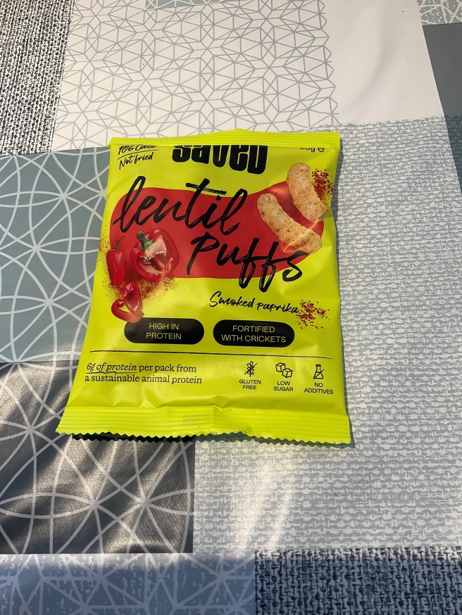A packet of Saved Lentil Puffs lies on a tablecloth with duck egg colour scheme patterns.