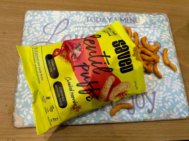 A packet of Saved Lentil puffs and some of the puffs lie on a blue placemat on a brown tabletop.