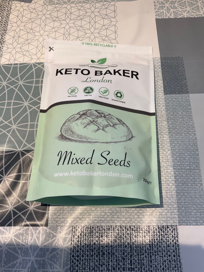 A packet of Keto Baker Mixed Seeds bread mix lies on a tablecloth with duck egg colour scheme patterns.