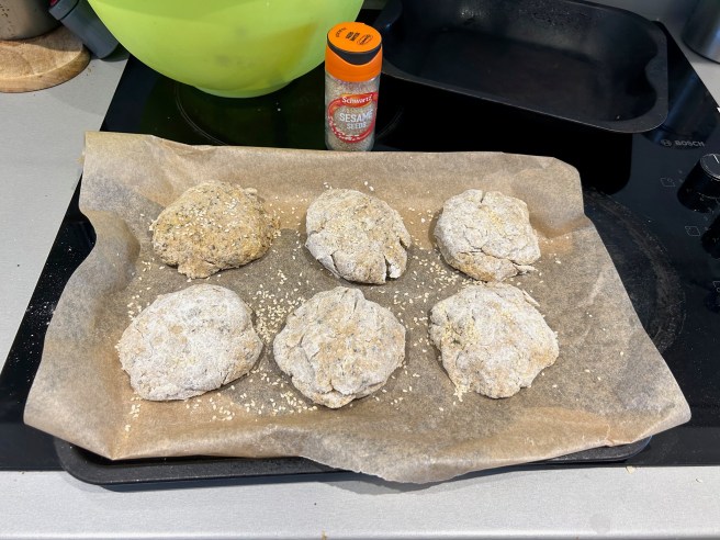 Some pre-baked bread rolls sit on a baking tray lined with brown greaseproof paper. A jar of sesame seeds stands behind the baking tray.