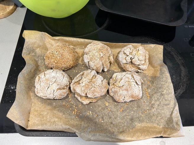 Some baked bread rolls sit on a baking tray lined with brown greaseproof paper.
