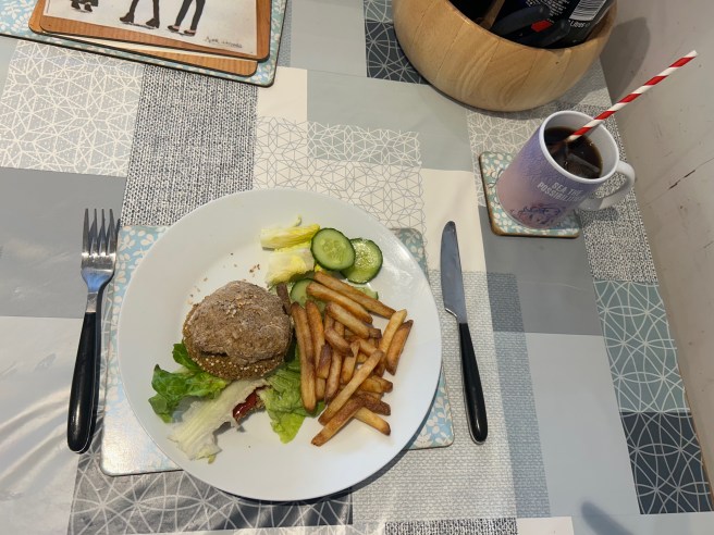 A plate of a vegan burger in a bread roll, French fries and salad sits on a tablecloth with duck egg colour scheme patterns.