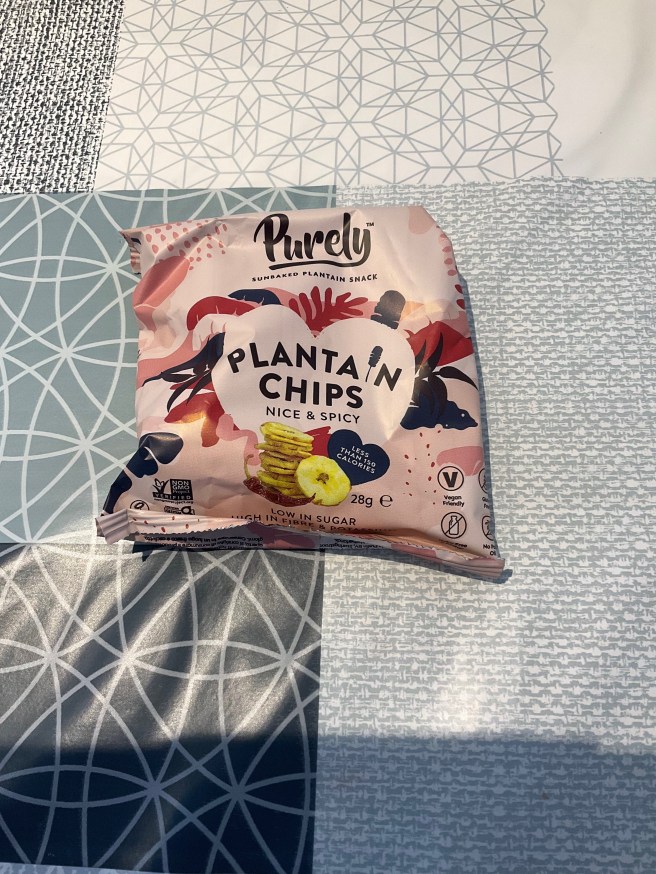 A packet of Purely Plantain Chips lies on a tablecloth with duck egg colour scheme patterns.