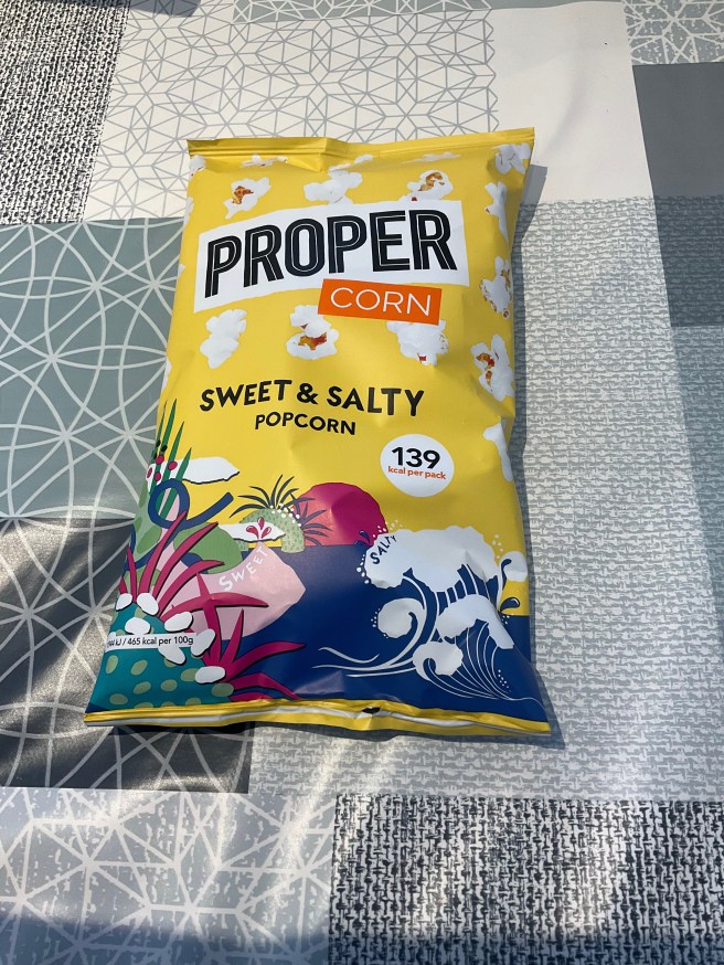 A packet of Proper Corn Sweet & Salty Popcorn lies on a tablecloth with duck egg colour scheme patterns.