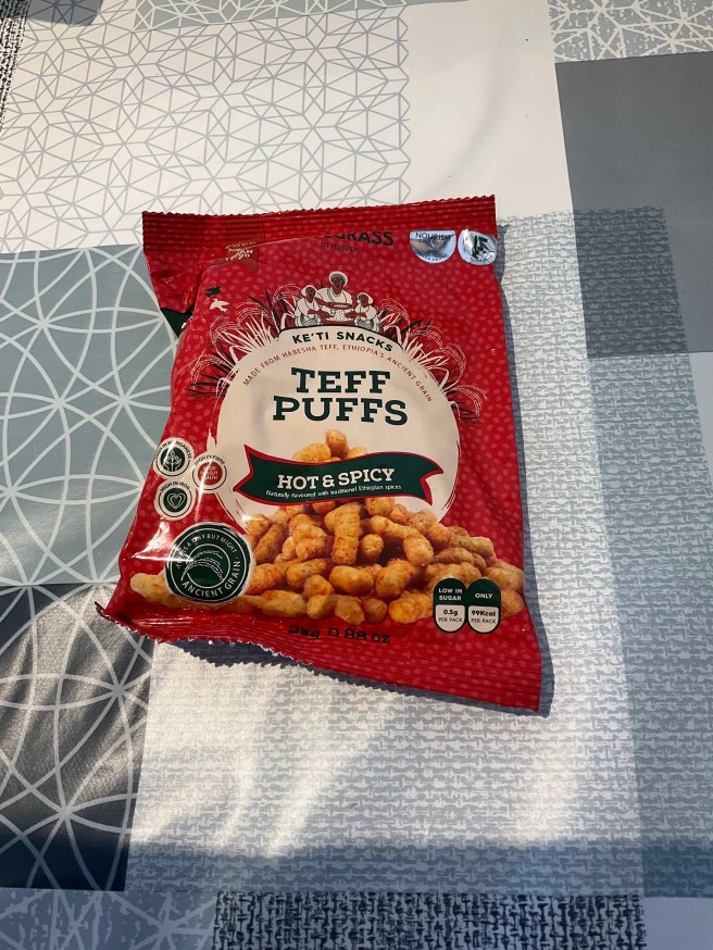 A packet of Lovegrass Teff Puffs lies on a tablecloth with duck egg colour scheme patterns.