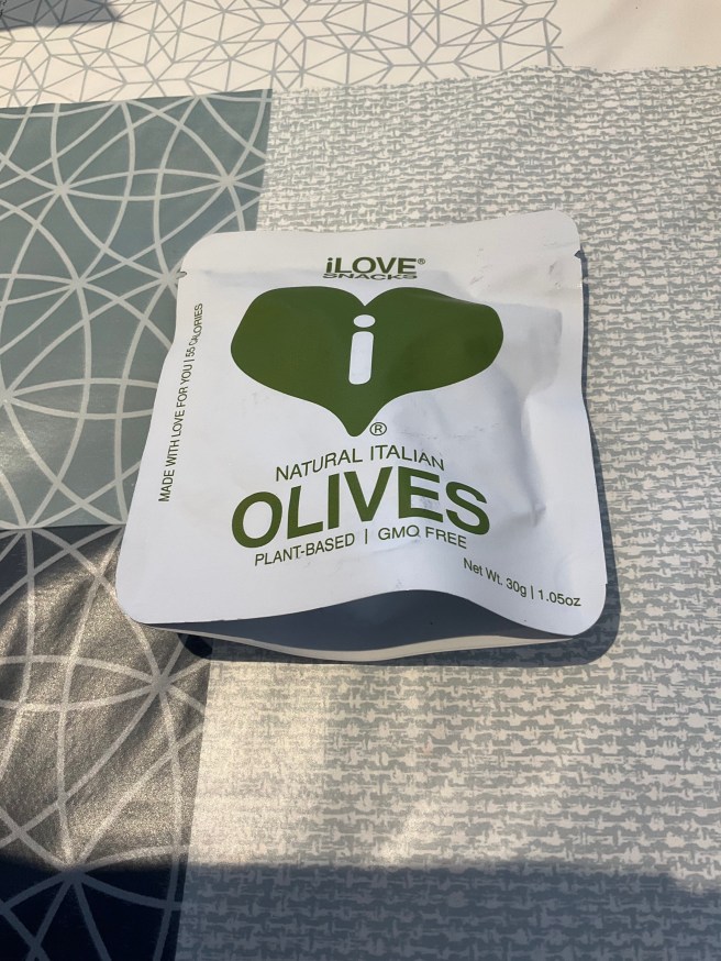A packet of iLove Snacks Olives lies on a tablecloth with duck egg colour scheme patterns.