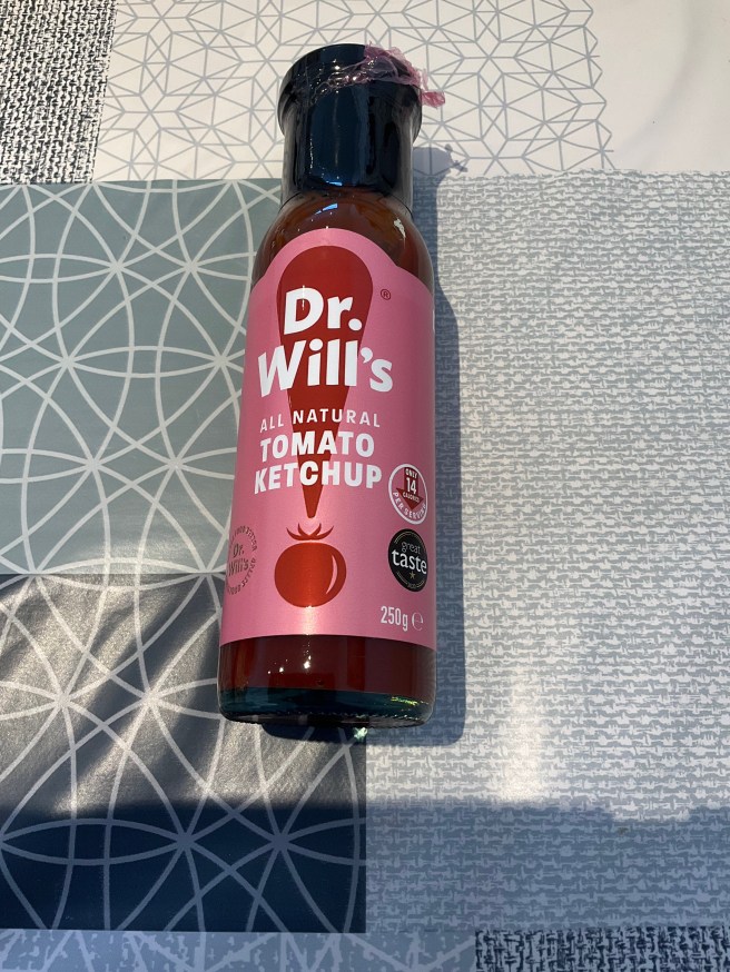 A glass bottle of Dr. Will's Tomato Ketchup lies on a tablecloth with duck egg colour scheme patterns.