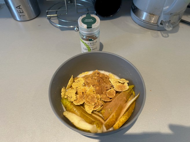 A bowl of Mesa Sunrise Cereal, some coconut milk, a pear, some peanut butter and some cinnamon sits on a grey kitchen worktop. A jar of cinnamon stands behind the bowl.