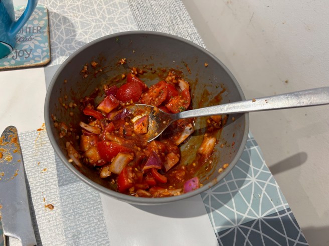 A bowl of salsa with a spoon in it stands against a white wall and on a table cloth with duck egg colour scheme patterns.