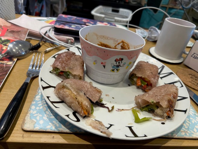 A plate of rice wrap spring rolls and a bowl of peanut butter dipping sauce sit on a blue placemat on a brown coffee table.