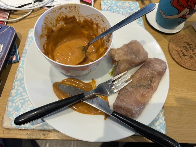 A plate of rice wrap spring rolls and a bowl of peanut butter dipping sauce sit on a blue placemat on a brown coffee table.