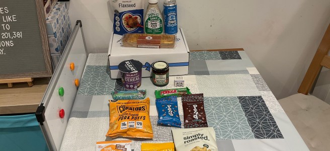 Various gluten-free food and drink products and a cardboard box lie on a duck patterned tablecloth. Some products lie on the cardboard box.